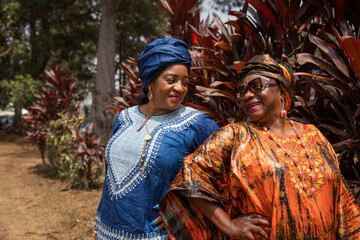 Two African female friends dressed in traditional clothing look at each other smiling during a walk.