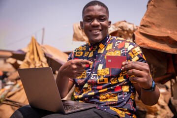 A boy makes purchases online with his credit card while using his laptop in an African market.