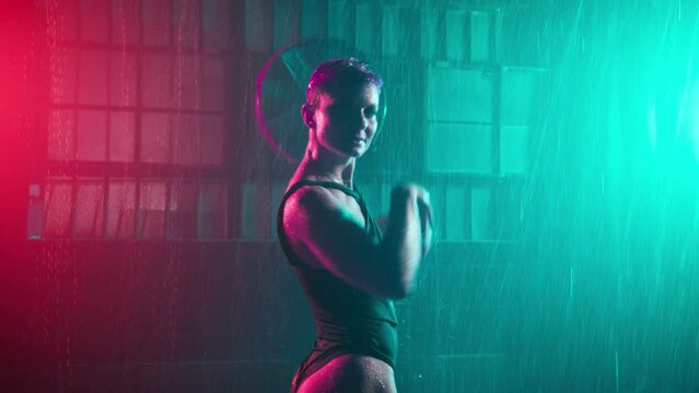 Stylish young girl dancing in grunge urban space, moving inside abandoned garage building under falling rain. Colorful studio spotlights. Half naked dancer on wet floor in futuristic cyberpunk space