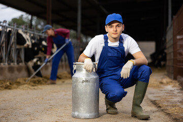 Portrait of young European man dairy farmworker in uniform carrying large metal milk can in cowshed
