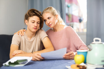 Caring mother calming her upset teenage son student receiving test failure notification while sitting at table in cozy dining room