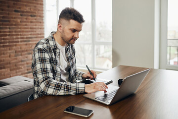 A professional photographer is sitting at his office desk. Man in office with digital graphics tablet and pen for drawing. A camera, a laptop, a card reader are all around on the table.