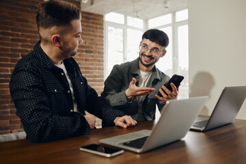 Fototapeta premium Two happy young men are working in the office on laptops. One shows the other something on the phone.