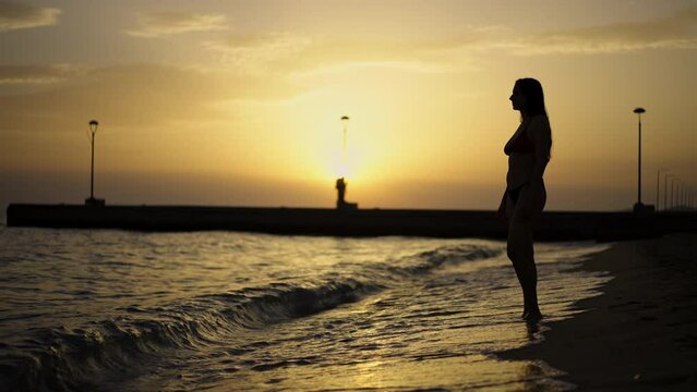 Woman in bikini standing on the beach and watching sea waves coming onto the beach in slow-motion at sunset