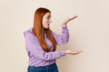 Young red hair woman isolated shocked and amazed holding a copy space between hands.
