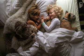 two sisters and dogs laying in bed holding hands giggling morning time