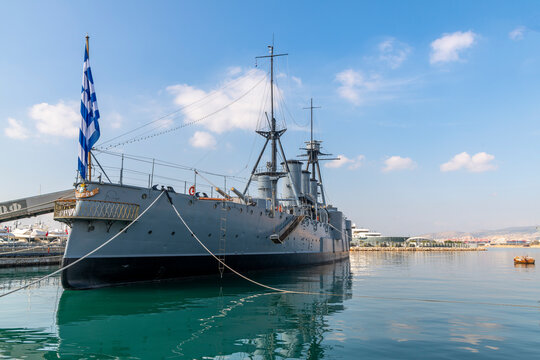 Oceanfront view of the Royal Hellenic Navy museum ship Averof in the Marina Flisvos harbor along the coast of Paleo Faliro, Greece, with the port city of Piraeus in the distance.