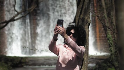 Young woman sitting in front of waterfall making selfie photos and enjoying the natural and peaceful view. Hiker check gps coordinates on smartphone app