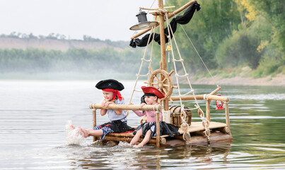 Children in pirate costumes play on a wooden raft at sunset. Two girls pretend to be the captains of a ship with black sails and a flag. Funny kids dreaming about adventure and travel. - obrazy, fototapety, plakaty