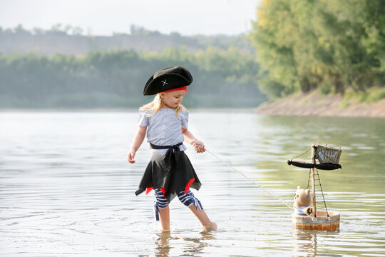 Children in pirate costumes play on a wooden raft at sunset. Two girls pretend to be the captains of a ship with black sails and a flag. Funny kids dreaming about adventure and travel.