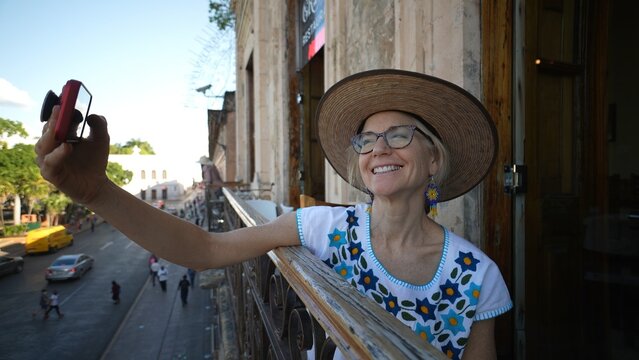 Wide angle view of pretty mature elderly woman wearing straw hat and glasses taking a selfie while on balcony over a public square in Europe or Latin America. Concept of digital nomad. - Powered by Adobe
