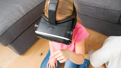 Young girl in VR headset touching air during virtual reality experience at home. Emotional kid...