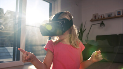 Young girl in VR headset touching air during virtual reality experience at home. Emotional kid...