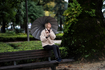 Fototapeta premium A woman with an open umbrella sits on the back of a bench in the Park.