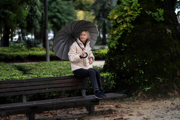 A woman with an umbrella sits on the back of a bench in the Park.