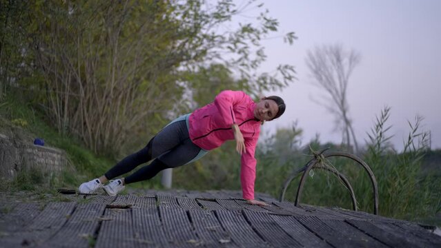 Attractive sporty girl exercising in slow-motion at riverside. Beautiful athletic young woman doing push ups near river at sunset