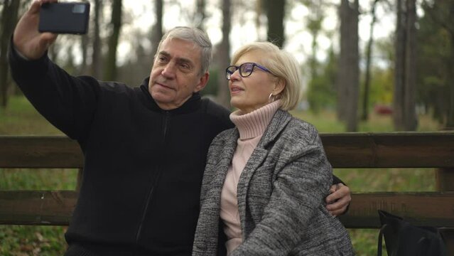 Happy smiling senior couple taking selfie on smartphone while sitting on park bench in the forest