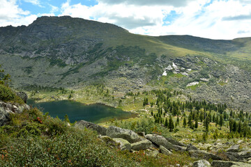 A view from the top of a rocky mountain to a small lake lying in a hollow surrounded by mountains on a cloudy summer day.