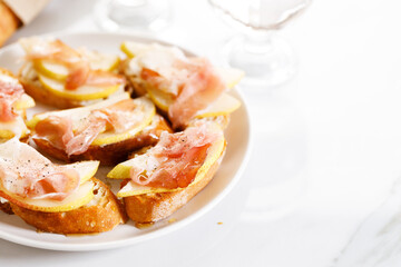 Crostini with cheese, sliced pear and prosciutto on white plate.