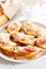 Crostini with cheese, sliced pear and prosciutto on white plate.