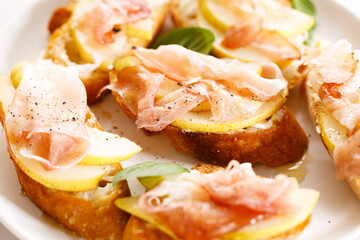 Crostini with cheese, sliced pear and prosciutto on white plate.