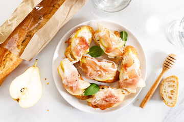 Crostini with cheese, sliced pear and prosciutto on white plate.