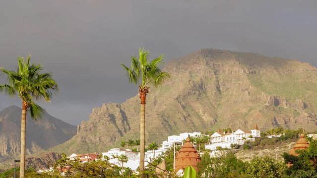 Beautiful timelapse of fast moving clouds at Las Americas (Tenerife, Spain), distant mountains, golden hour light before the sunset, palm trees in foreground, wide shot
