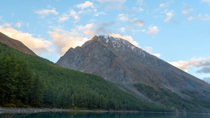 The shore of the turquoise lake Shavlinsky with a forest surrounded by rocks and a forest with snow in Altai during the day.