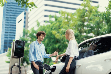 Progressive businessman and businesswoman with electric car parking and connected to public...