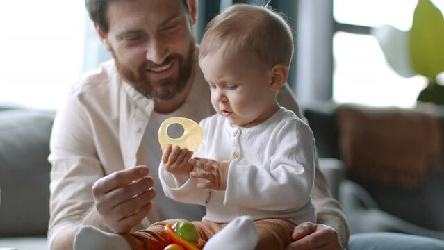 Happy childhood. Carefree little baby girl playing with toy and teether at home, loving dad sitting near and laughing