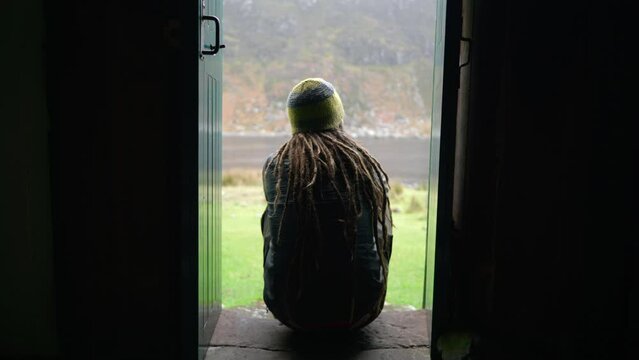 A man with dreadlocks and wearing hiking gear sits on a step in a narrow doorway of a bothy in the Highlands of Scotland to enjoy the view outdoors.