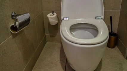 White ceramic toilet bowl with the lid raised. The walls and floor of the  restroom are lined with beige tiles. Rolls of toilet paper, a washing brush are visible.