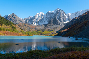 The shore of the alpine lake Shavlinskoye in Altai in the shade against the backdrop of sun-drenched peaks with snow and glaciers. Mountains Dream, Fairy Tale, Beauty.