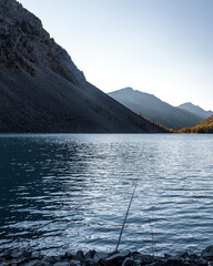 A fishing rod lies in the shade on the rocks on the shore of the alpine lake Shavlinskoe with sunlight in the mountains on the horizon. Vertical frame.