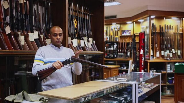 Portrait of hispanic salesman showing collectible old rifled musket on background with rack full of vintage and modern firearms in gun store 