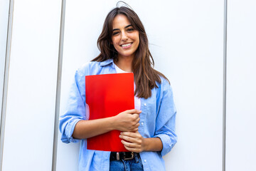 Happy, smiling female college student looking at camera holding red folder.