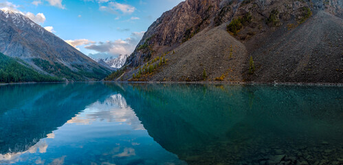 Panorama of the turquoise lake Shavlinskoe in the shade with stones among the mountains with reflection of the peaks with glaciers and snow in Altai.