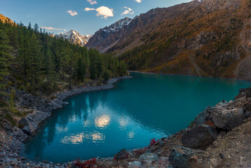 The bay of the mountain lake Shavlinskoye in Altai with stone shores in the shade with snowy mountains in the sun.