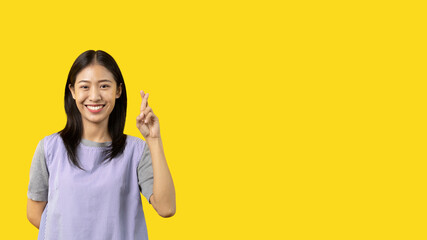 Young Asian woman making a symbolic gesture with fingers crossed showing good luck, White lie gesture, Fingers crossed, Woman doing hand sign on yellow background, Superstitious concept.