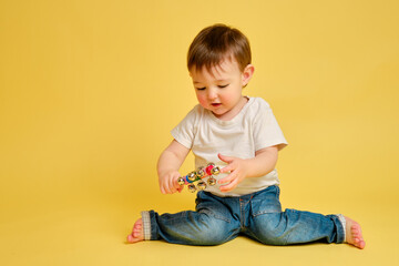 Toddler baby plays with a musical rattle bells on a stick, studio yellow background. Happy child musician playing percussion musical instrument, copy space. Kid aged one year and four months - obrazy, fototapety, plakaty