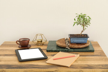 Table with Bonsai tree, tablet computer, cup of beverage and notebook near light wall