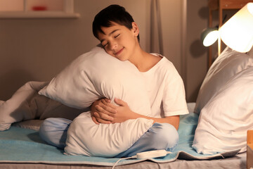 Little boy with pillow sitting on electric heating pad in bedroom at night