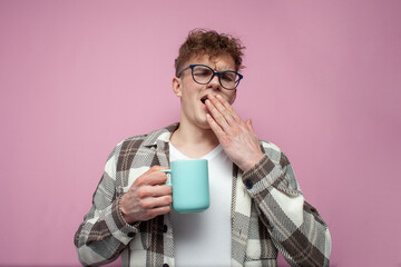 tired sleepy guy in glasses holds a mug of coffee and yawns on pink background, awake student with...