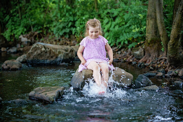 Fototapeta na wymiar Cute little toddler girl having fun by a river on warm and sunny summer day. Happy excited preschool child splashing with water in forest stream creek.