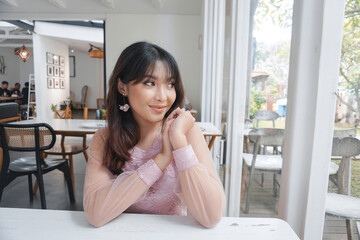 A smiling Asian woman wearing pink dress while sitting near window