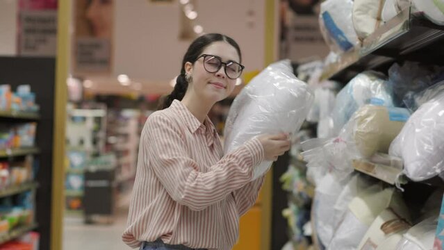 Portrait of young Caucasian woman wearing eyeglasses choosing pillow for home and family. Shopping in furniture store.