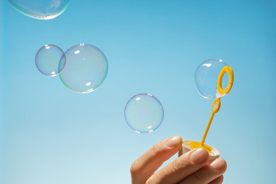 Close up side view shot of young man blowing soap bubbles on blue background. Focus on hands and wand.
