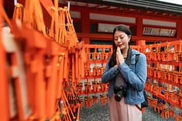 selective focus of pious asian Japanese girl surrounded by red wood plaques ema and making wish with hands together at Fushimi Inari Taisha shrine in Kyoto japan - obrazy, fototapety, plakaty