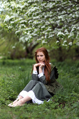 Young red haired girl in green vintage dress in blooming garden. Romantic portrait of old fashion dressed woman sitting on grass