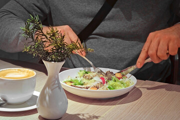 Young Man Wearing Gray Sweater Eating Fresh Vegetable Salad. Unrecognized Male Person Eating Caesar Salad in a Cafe or Restaurant and drinking Coffee while Sitting at a Wooden Table. Business Lunch.